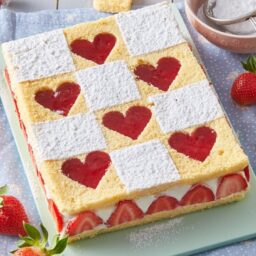 Prăjitură cu inimioare de căpșuni și cremă de mascarpone pe un suport gri, alături de căpșuni proaspete, un bol cu sită și zahăr și decupaje în formă de inimioare