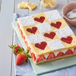 Prăjitură cu inimioare de căpșuni și cremă de mascarpone pe un suport gri, alături de căpșuni proaspete, un bol cu sită și zahăr și decupaje în formă de inimioare