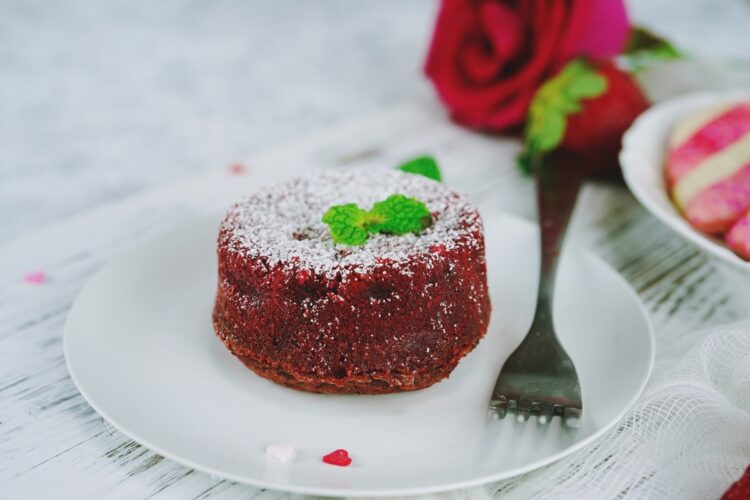 Red Velvet Lava Cake pe o farfurie, decorată cu zahăr pudră și frunze de mentă, alături de o furculiță și un trandafir roșu