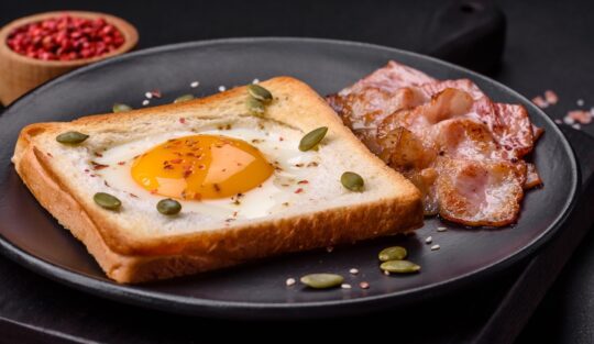 Ou prăjit în pâine și decorat cu semințe pe o farfurie neagră, alături de felii de bacon crocante