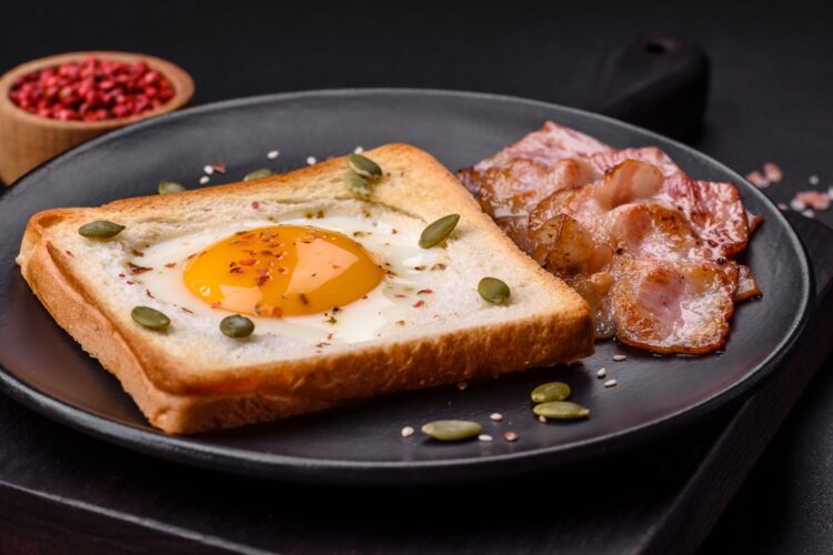 Ou prăjit în pâine și decorat cu semințe pe o farfurie neagră, alături de felii de bacon crocante
