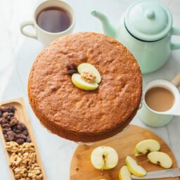 Prăjitură cu mere și nuci, aromată cu scorțișoară pe un platou, alături de un ceainic, o ceașcpă cu ceai, un platou cu nuci și stafide , mere feliate și un ibric cu cafea