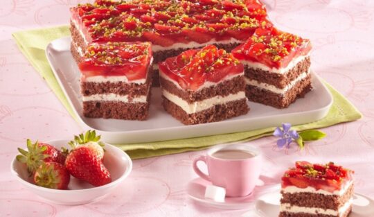 Porții de prăjitură cu căpșuni și fistic pe un platou, o porție pe o farfurie, o ceașcă cu cafea și un bol cu căpșuni
